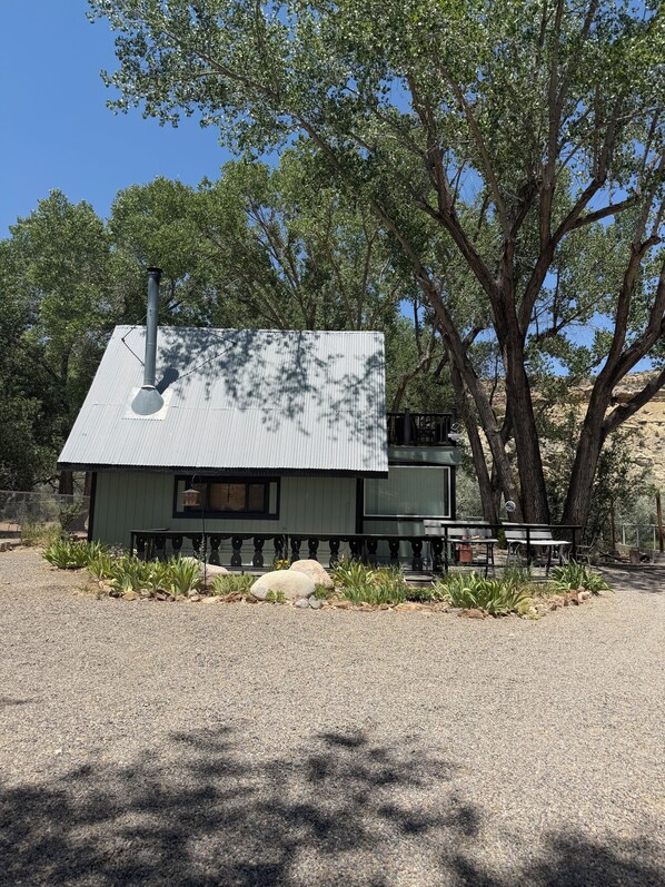 A small, single-story building with a metal roof and a chimney, surrounded by gravel and plants, with trees in the background. - Spectacular Cabin (Navajo Dam)