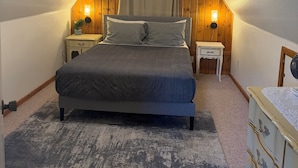 Un dormitorio con techo inclinado, una cama, mesitas de noche con lámparas, un aparador y una ventana con cortinas.