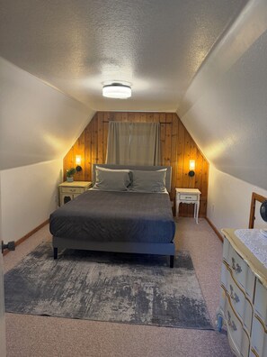 A bedroom with a sloped ceiling, a bed, bedside tables with lamps, a dresser, and a window with curtains.