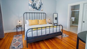 Een slaapkamer met een bed met metalen frame, een groot abstract schilderij, een nachtkastje met een lamp en een houten vloer.