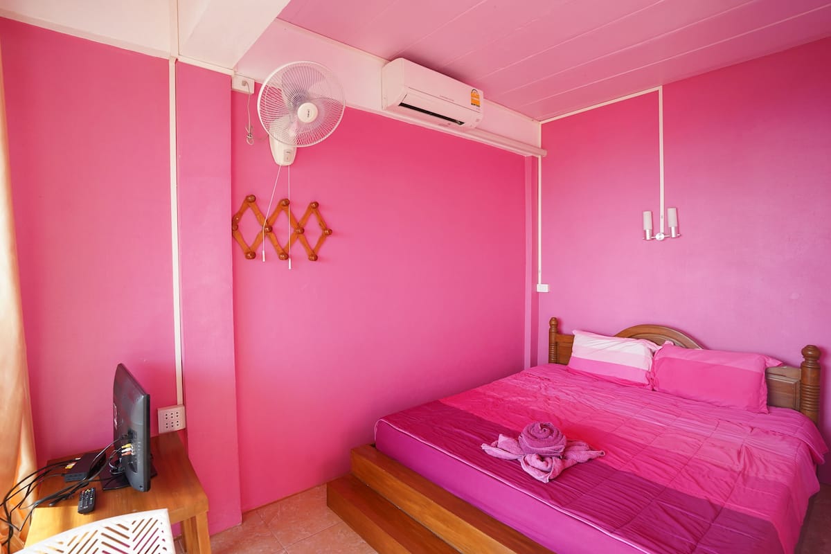 Una habitació amb decoració rosa, un llit, un televisor i un ventilador.