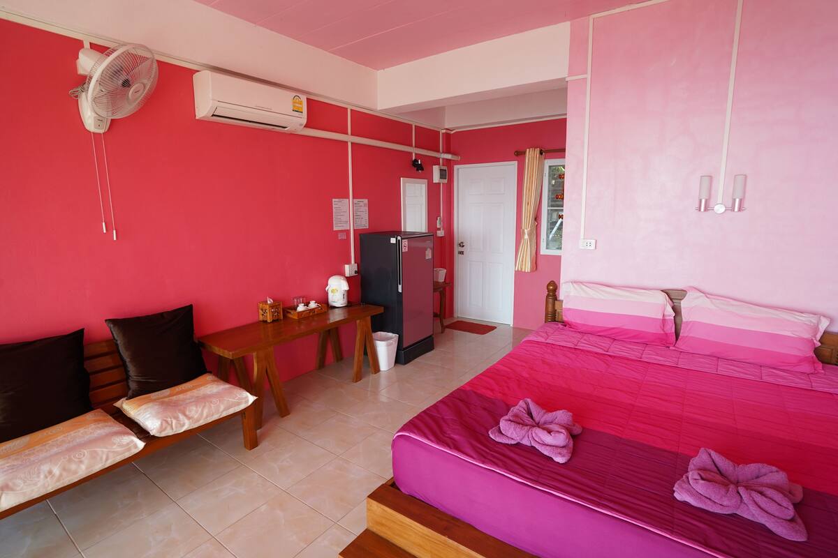 Una habitació amb llit, escriptori, cadira, ventilador i nevera.