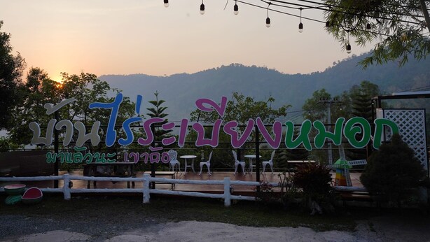 A sign with Thai characters, a wooden fence, and outdoor seating.
