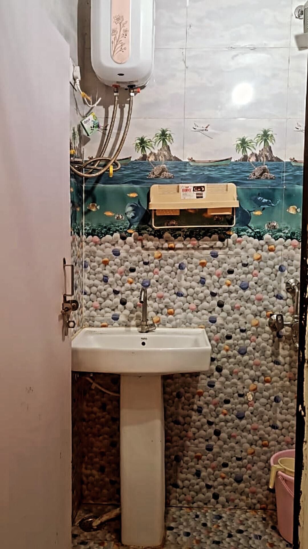 Une salle de bain avec un lavabo blanc, un climatiseur fixé au mur et un mur recouvert de carreaux orné d’une fresque aux motifs tropicaux.
