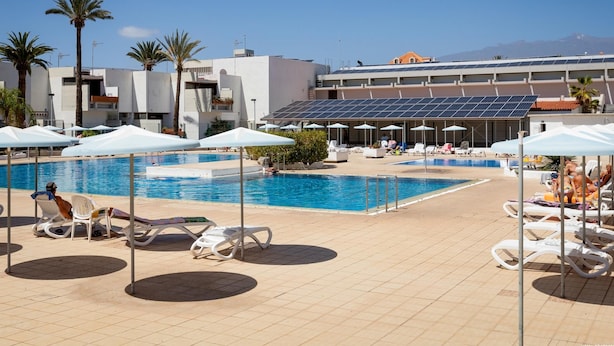 A pool area with sun loungers and umbrellas, a clear sky, and a building with solar panels in the background. - 535 Primavera Bright Accommodation With Pool (Costa del Silencio)