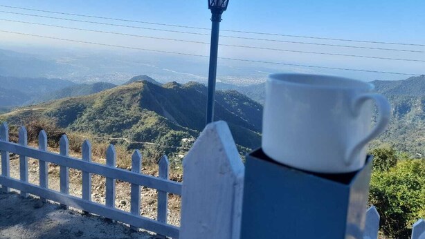 Una cerca blanca de listones, un poste de luz azul y una taza blanca sobre una mesa con vistas a un paisaje montañoso.