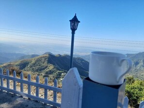 A white picket fence, a blue lamp post, and a white mug on a table with a view of a mountainous landscape. - Aranyam Hills (Lansdowne)