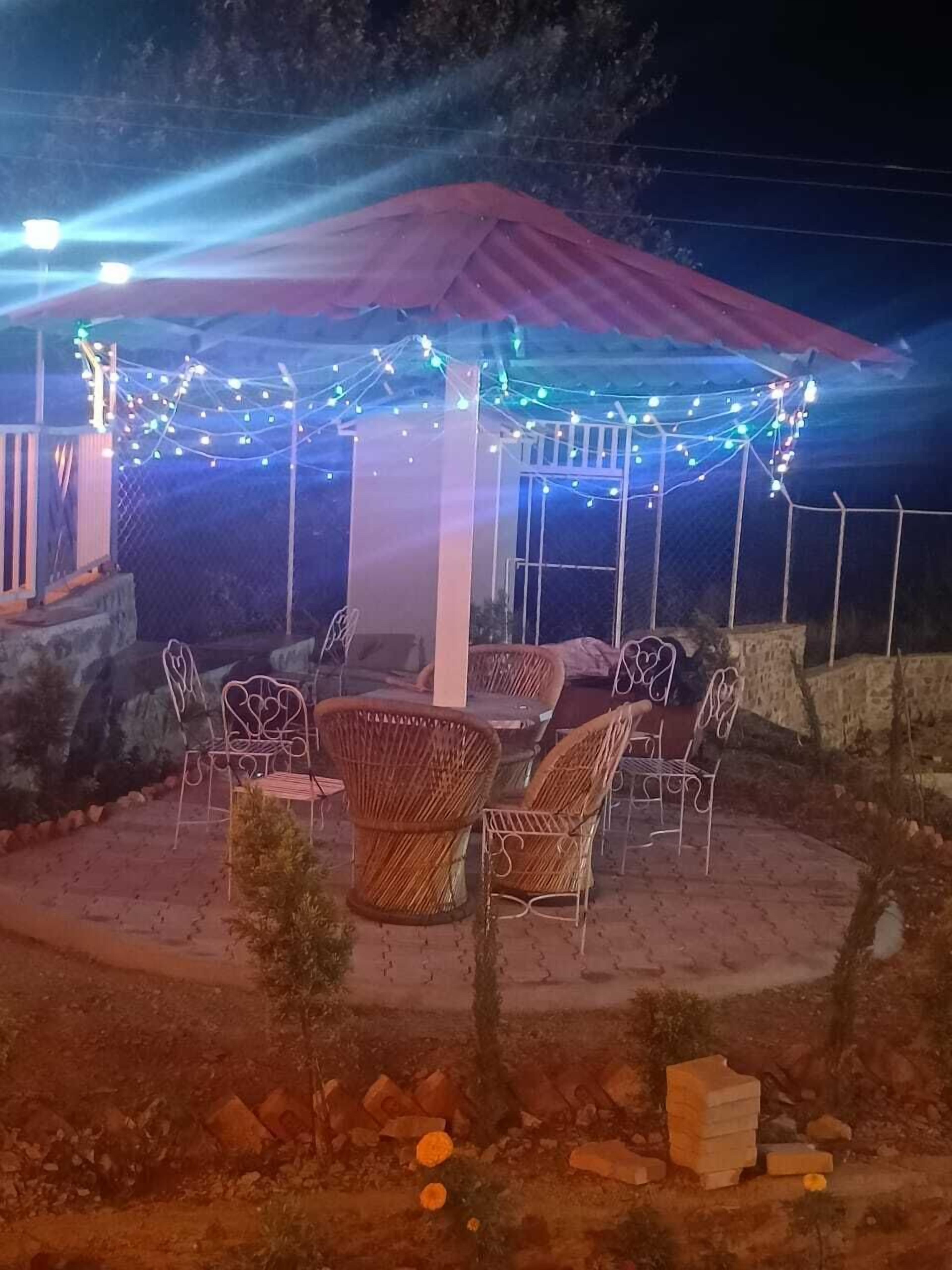 Un área de estar al aire libre con sillas de mimbre y una mesa, cubierta por un dosel rojo y luces colgantes.