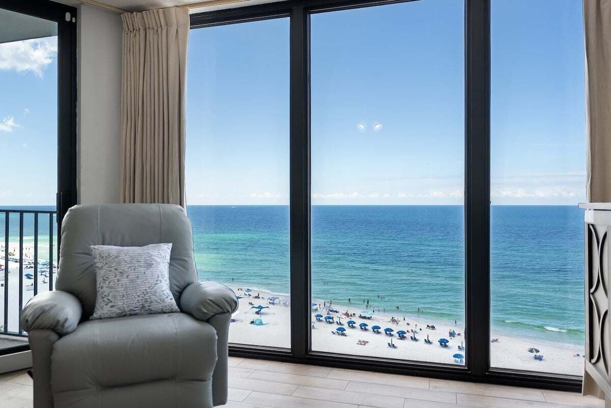 A balcony with a view of a beach, a grey armchair, and a large window.