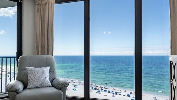 A balcony with a view of a beach, a grey armchair, and a large window. - Edgewater T2 #911- 2 BR + Hall Bunks! (Panama City Beach)