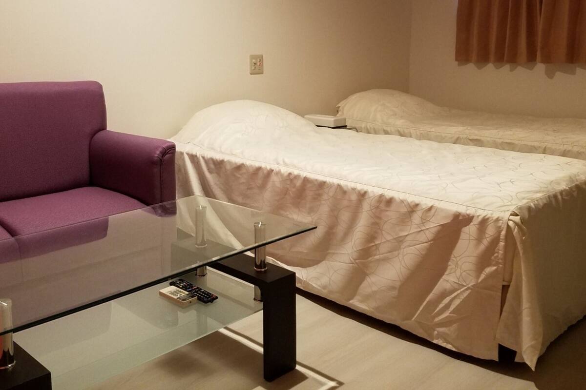 A small room with a glass table, a purple chair, and a bed with a white bedspread.