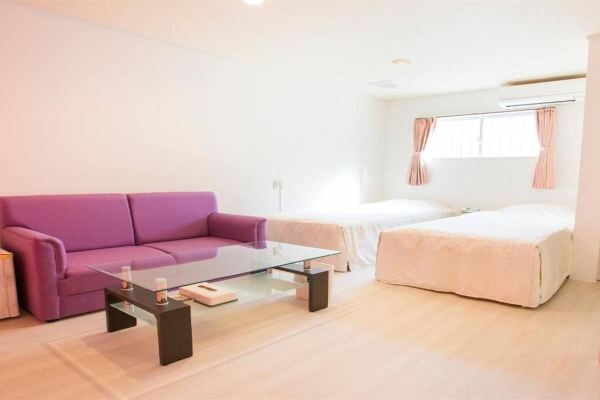 A modern room with a purple sofa, a glass coffee table, a bed with a white duvet, and a small kitchen area with a sink and countertop.