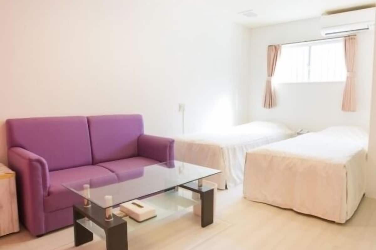 A modern room with a purple sofa, a glass coffee table, a bed with a white duvet, and a window with curtains.