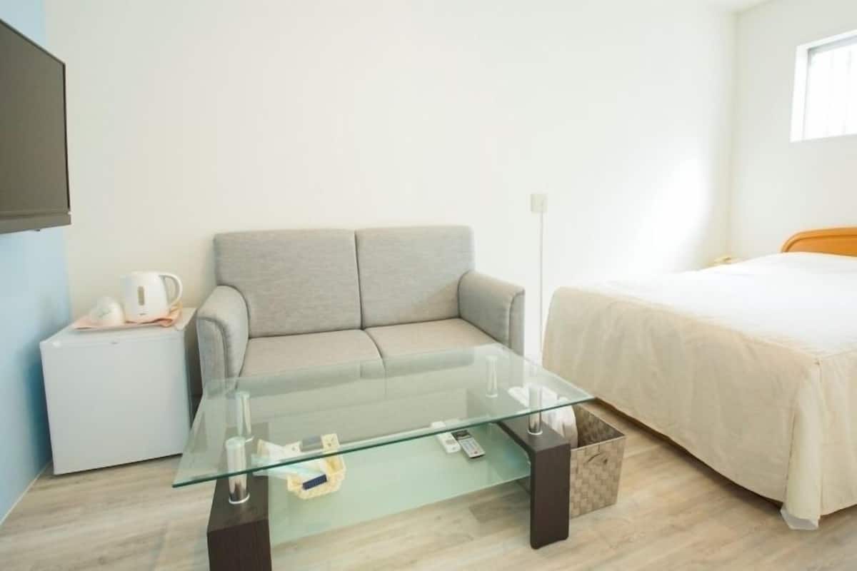 A room with a bed, a sofa, a glass coffee table, a small white table with a kettle, and a TV mounted on the wall.