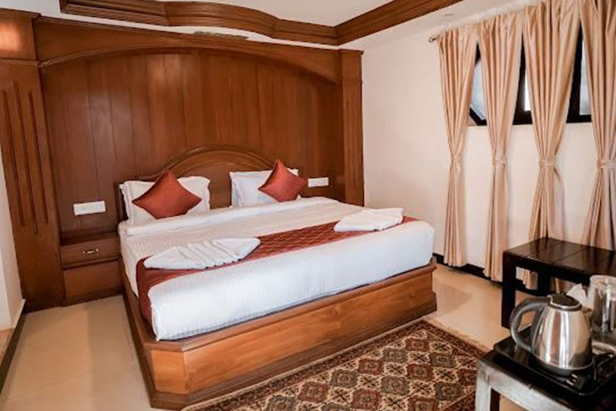 Una habitación de hotel con cabecera de madera, cama con ropa de cama blanca y almohadas rojas, una mesita con una tetera y una alfombra estampada.