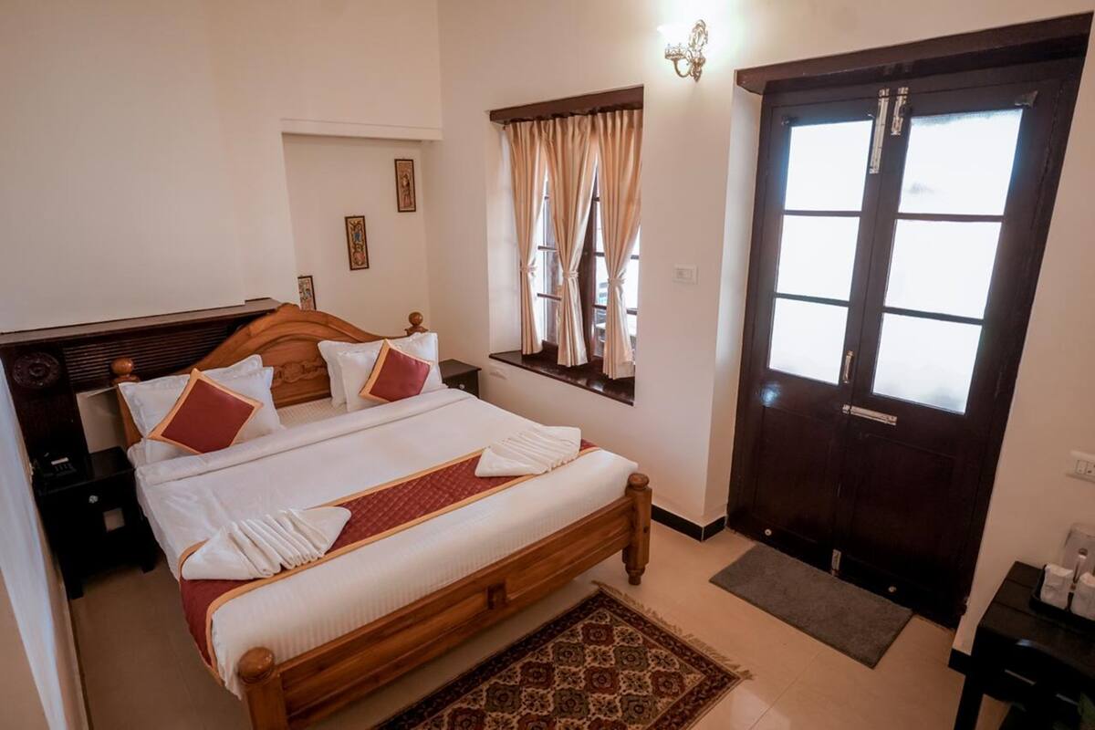 Una cama bien hecha con cabecera de madera, una ventana con cortinas, una puerta de madera oscura y una alfombra pequeña en el suelo.