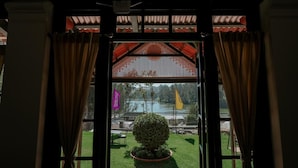 A view through a window with curtains, revealing a well-maintained lawn, a flag, and a body of water in the background. - Lakefront Home Hotel (Ootacamund)