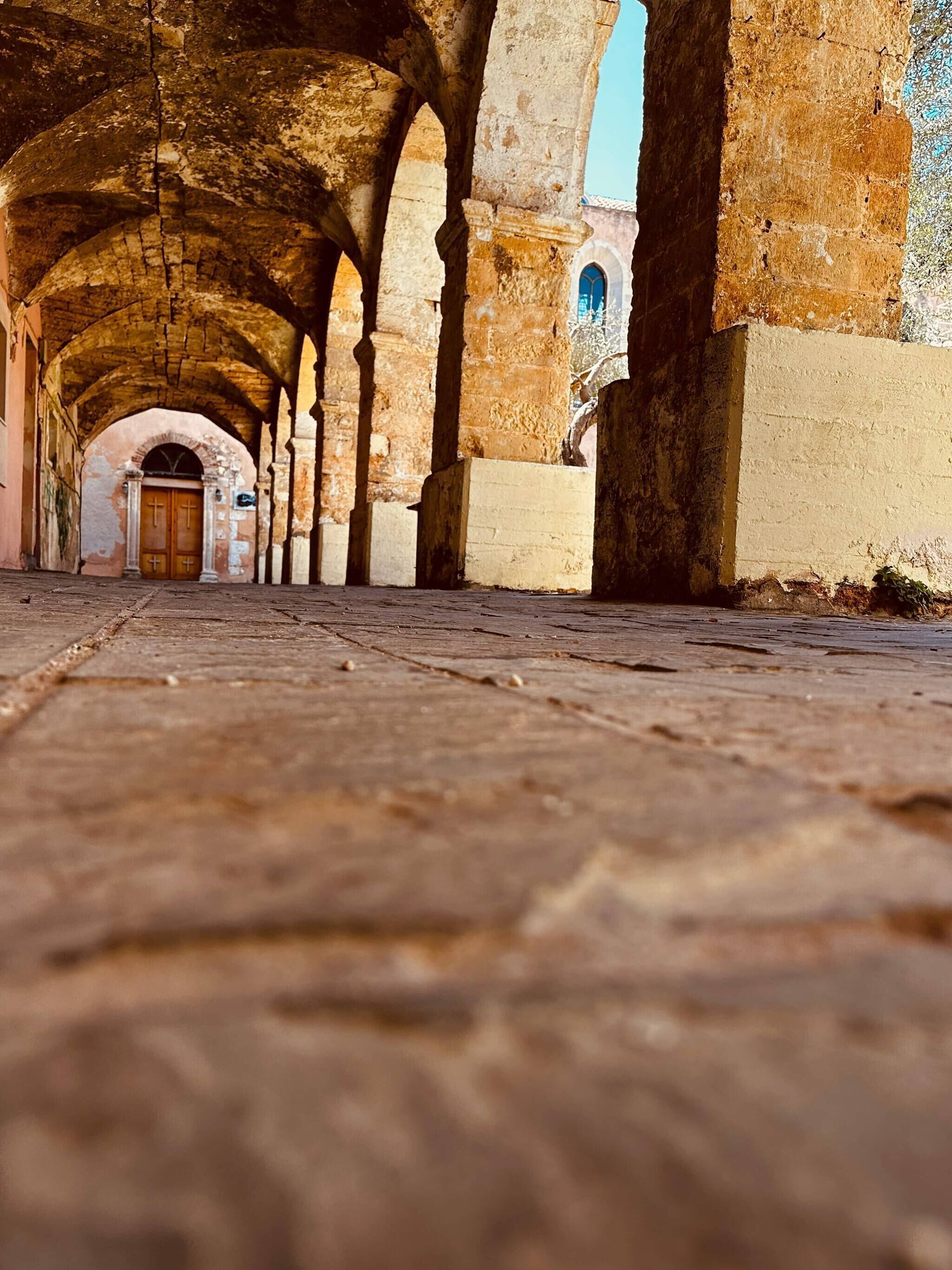 An arched corridor with stone columns, a wooden door at the end, and a wall with a window.