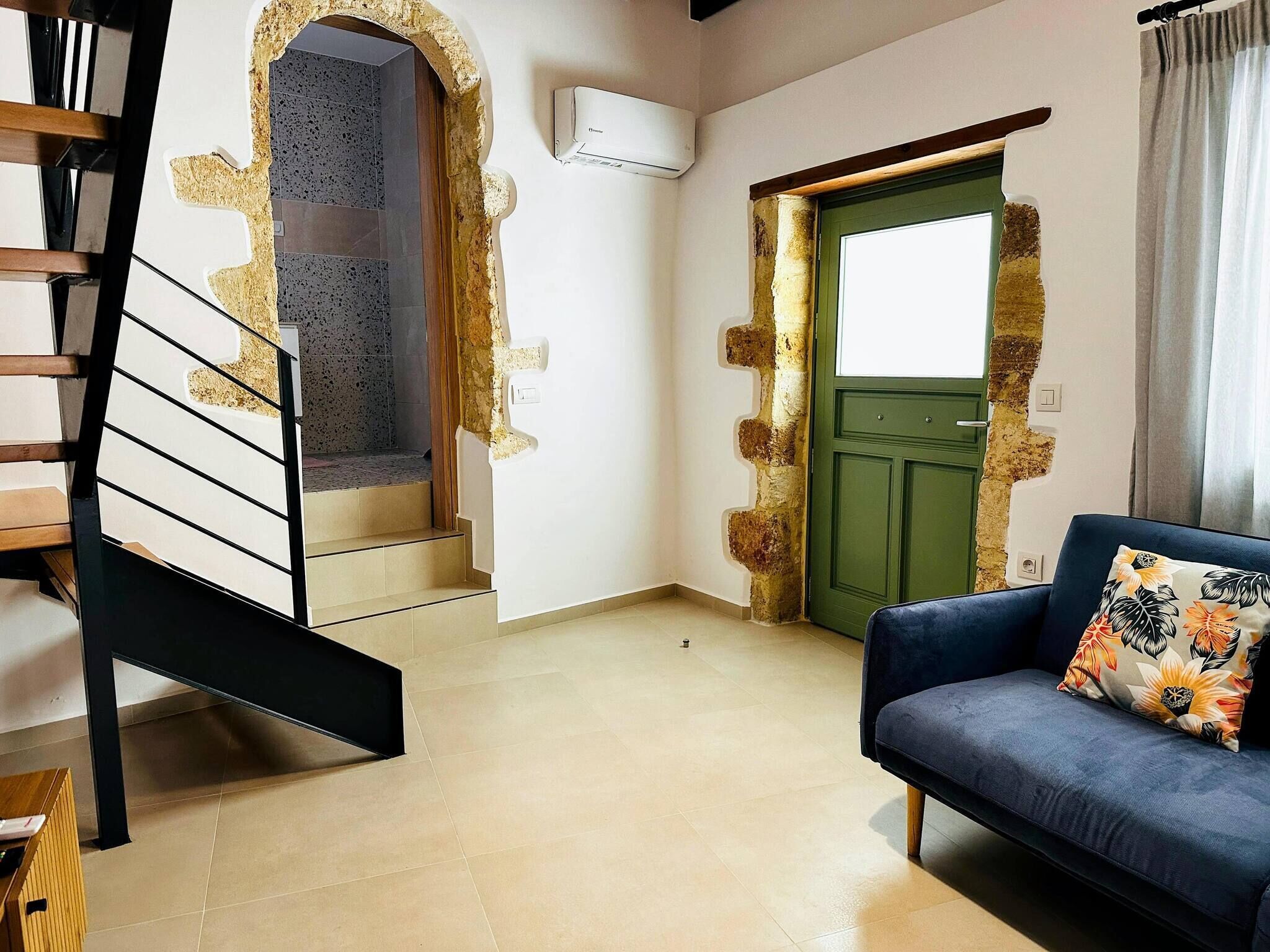 A staircase leading to a balcony, a green door, a blue sofa with a floral pillow, and a wall-mounted air conditioner.