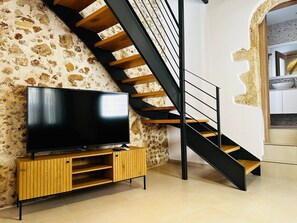 Un interior moderno con un televisor sobre un pedestal de madera, una escalera negra y una zona de cocina.