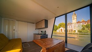 A modern room with a wooden table, a sofa, and a view of a historic building across a body of water. - Boat 'Hausboot Explorer' with Lake View and Private Terrace (Schwerin)