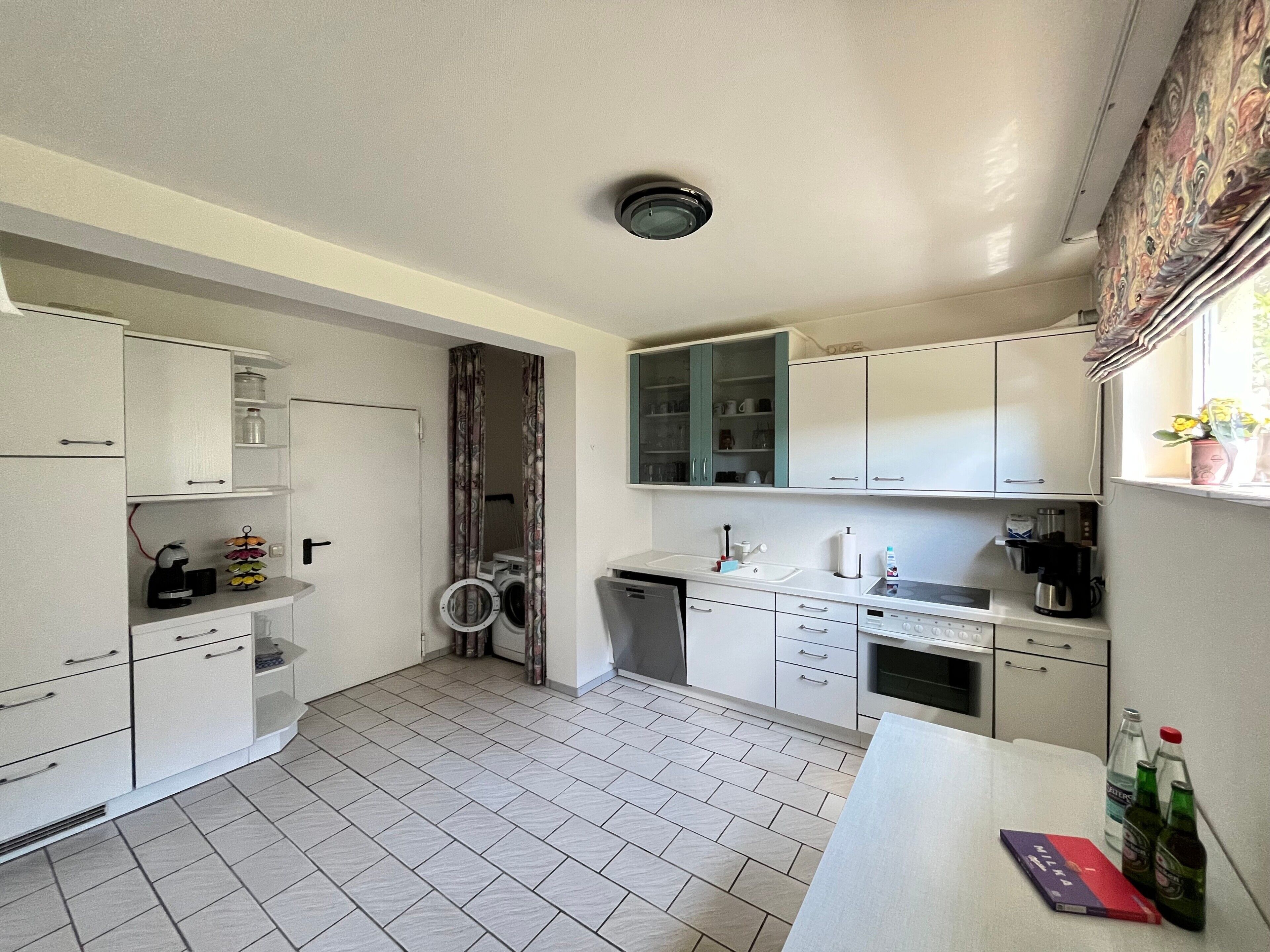 Cocina compacta con armarios blancos, lavadora, cafetera y una ventana con cortinas.