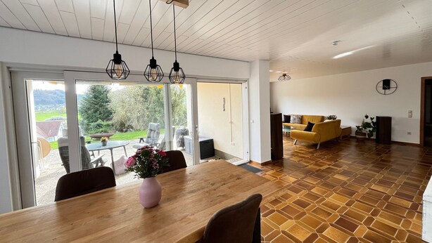 Un vaste espace de vie comprenant une table à manger en bois, un canapé jaune et une grande porte-fenêtre coulissante donnant sur un jardin.