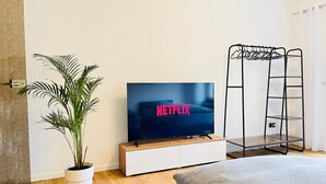 Une chambre à coucher moderne avec un téléviseur à écran plat affichant le logo de Netflix, une plante en pot, une table de chevet en bois, une armoire blanche, un canapé gris et un escabeau en métal noir.