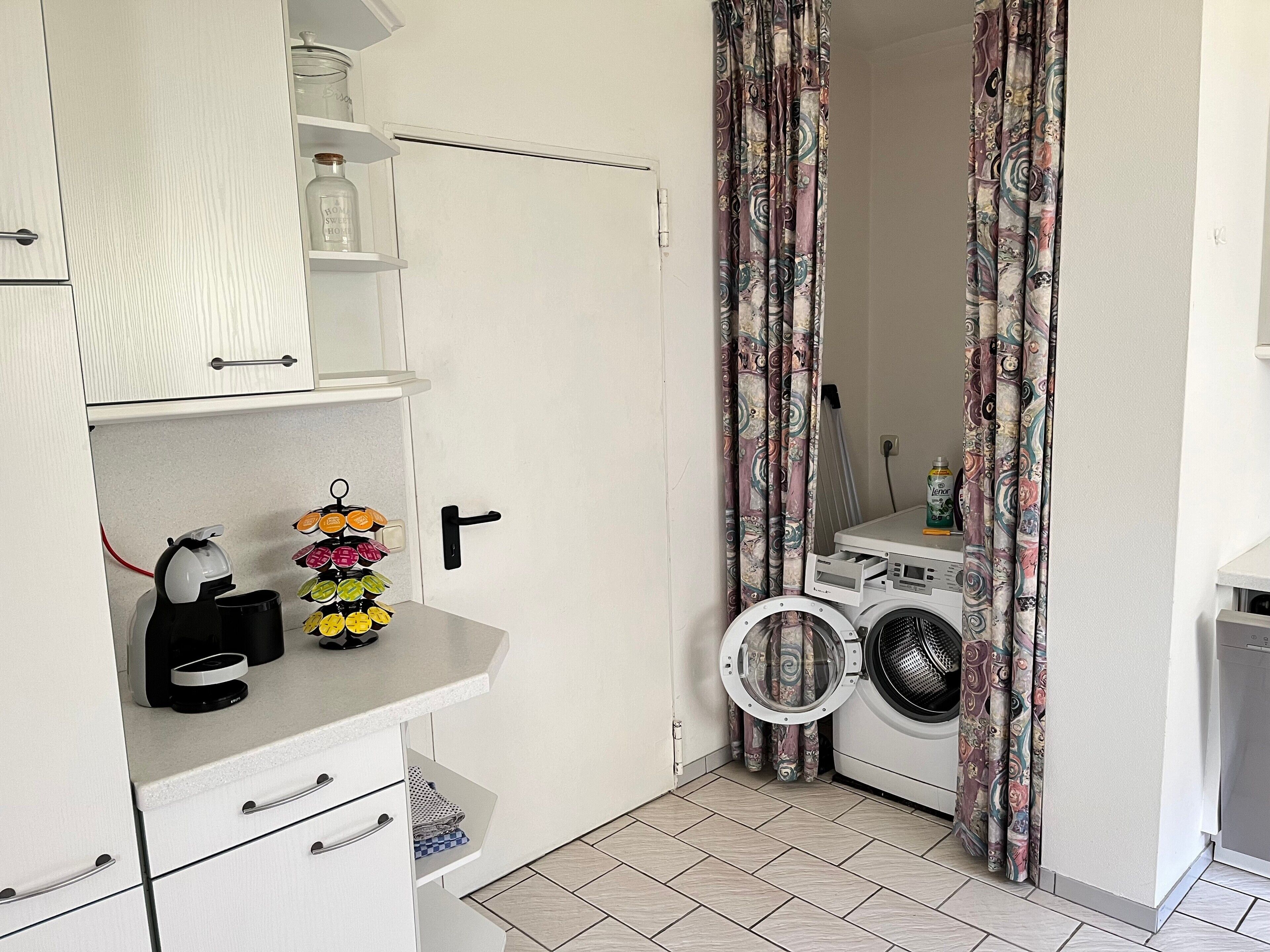 Une petite buanderie comprenant un lave-linge, un étendoir et une étagère avec des bocaux et une bouilloire.