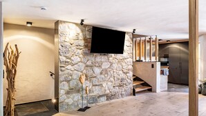 Un intérieur moderne avec un mur en pierre, un téléviseur fixé au mur, un escalier en bois et un coin cuisine.