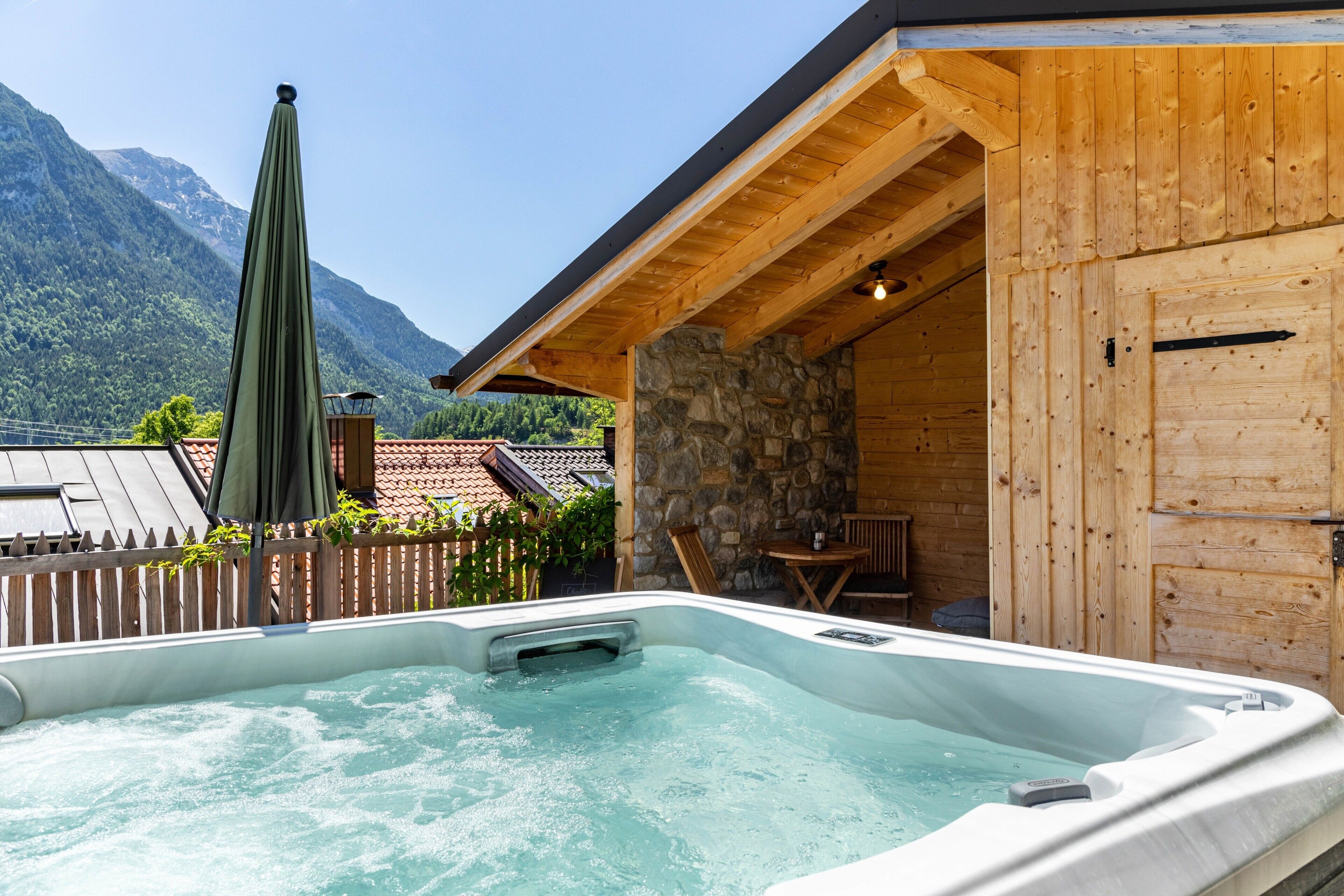 A hot tub with a wooden deck and stone wall, surrounded by a wooden fence, with mountains in the background.