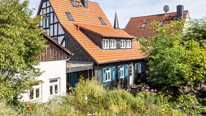 A traditional half-timbered house with a red-tiled roof, white walls, and blue shutters, surrounded by lush greenery.
