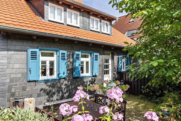 Une maison en pierre avec des volets bleus, un banc en bois et des fleurs roses dans le jardin.