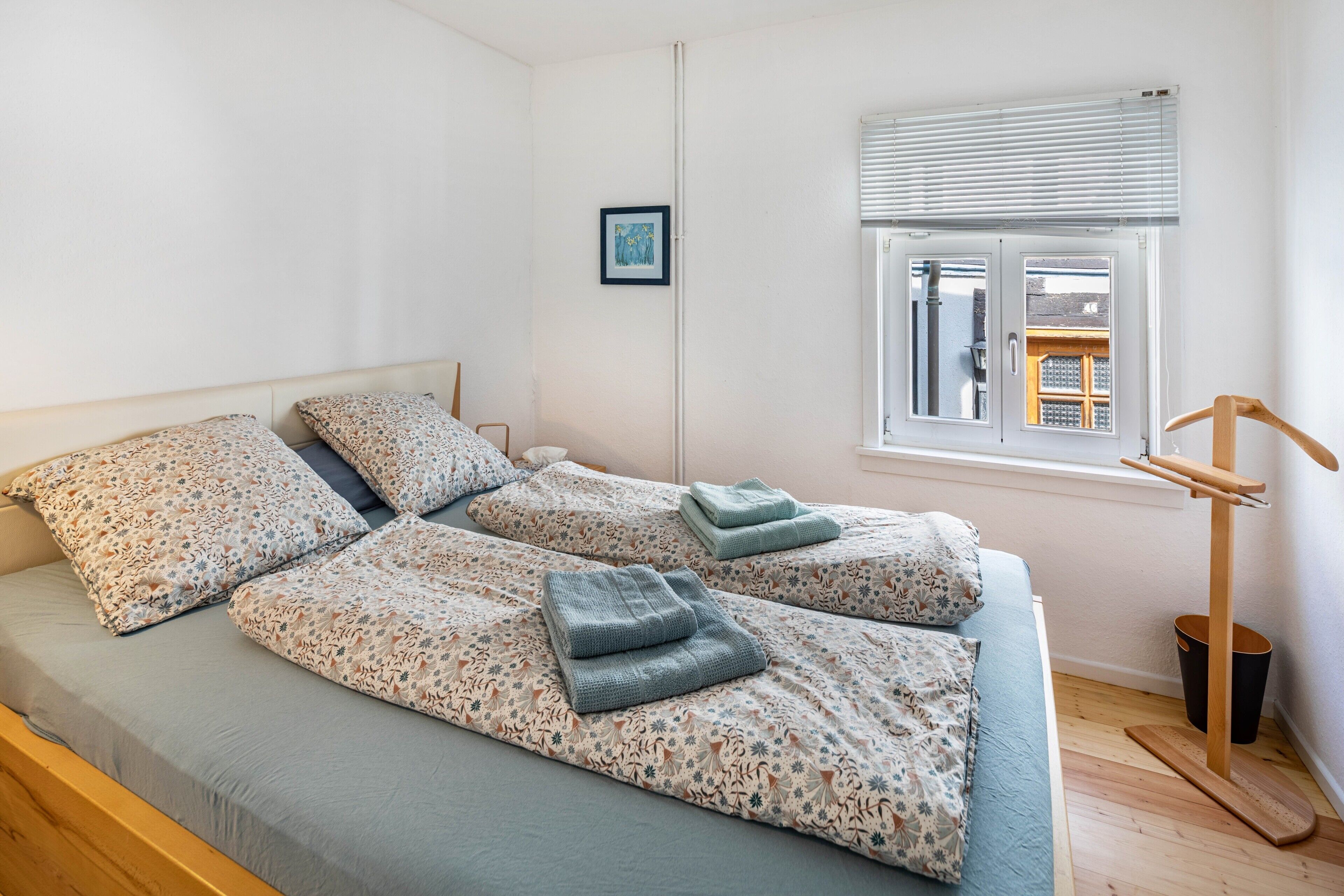 Una cama doble con ropa de cama floral, una ventana pequeña con persianas y suelo de madera.