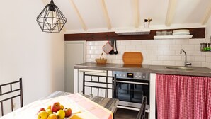 Une cuisine compacte avec une table à manger, un panier de fruits, un luminaire suspendu, une planche à découper en bois et un rideau à carreaux rouges et blancs.