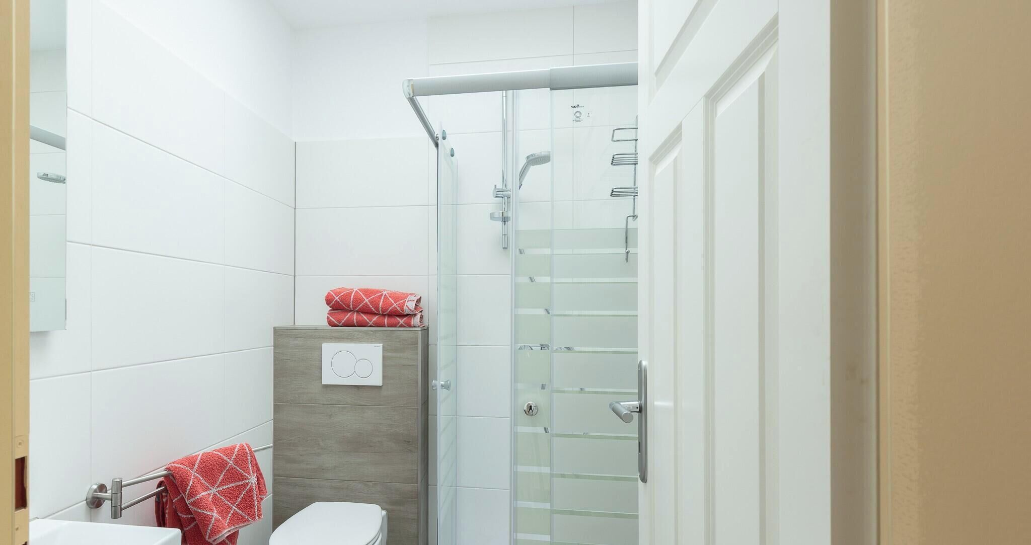 Un baño moderno con azulejos blancos, cabina de ducha de vidrio, un mueble de madera y un inodoro.