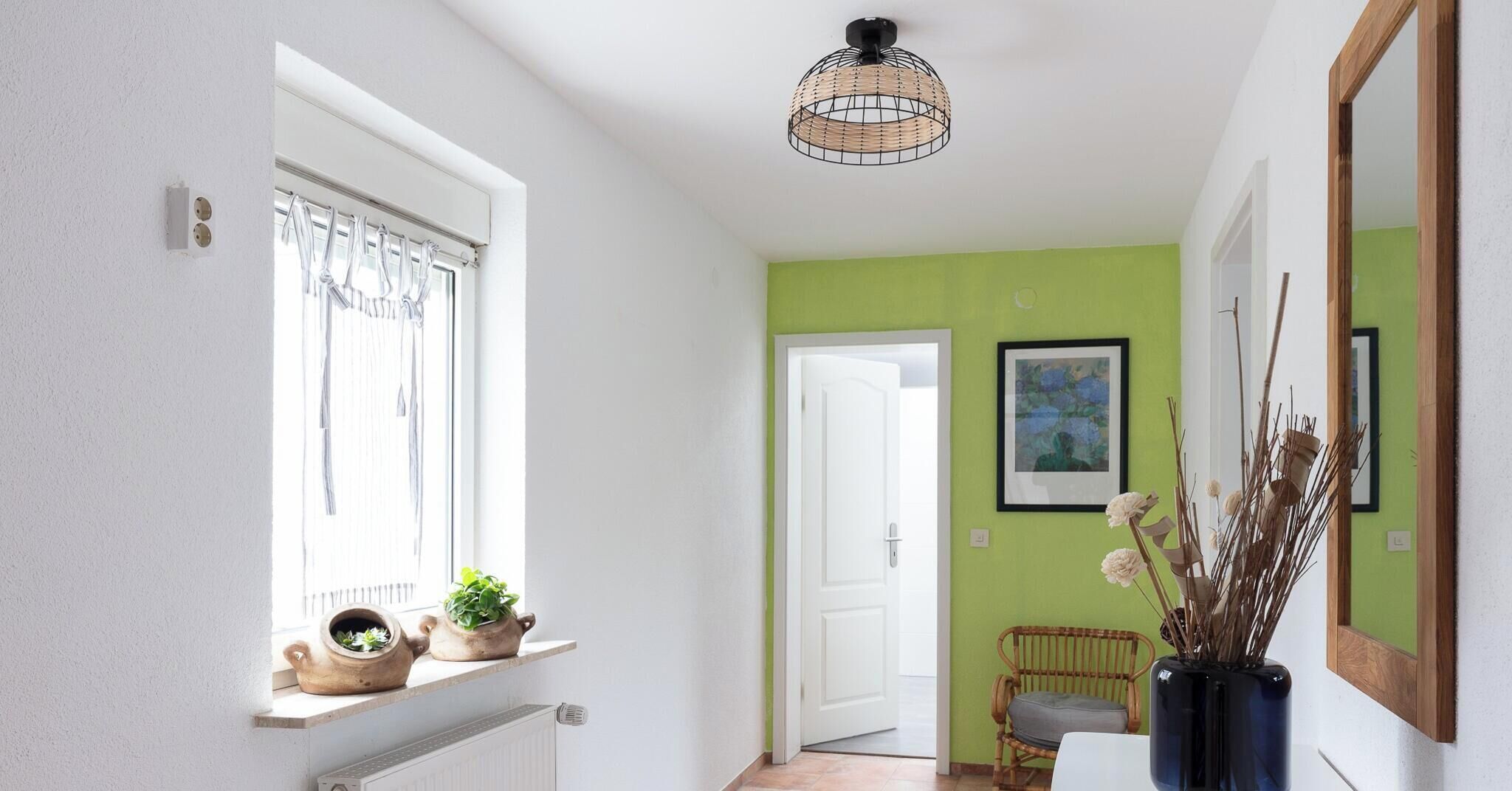 Un couloir avec une fenêtre, une porte, une chaise en osier et un vase avec des plantes.