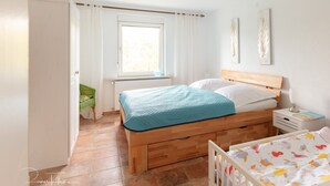 Ein Schlafzimmer mit einem hölzernen Bett, einem Nachttisch mit Lampe, einem Fenster mit Vorhängen und einem Babybett mit einem Bettlaken mit Vogelmuster.