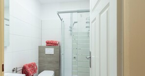 A modern bathroom with white tiles, a glass shower enclosure, a wooden cabinet, and a toilet.