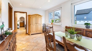 A dining area with a wooden table and chairs, a cabinet, and a window with a view of a house. - Apartment "Kebben" with Shared Terrace, Garden, and Pool (Kirchhundem)