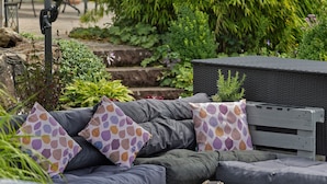 A garden setting with a grey outdoor sofa, patterned cushions, and a wicker basket. - Apartment "Kebben" with Shared Terrace, Garden, and Pool (Kirchhundem)