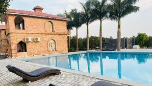 Una piscina con lettini, un edificio in mattoni con finestre ad arco e palme.