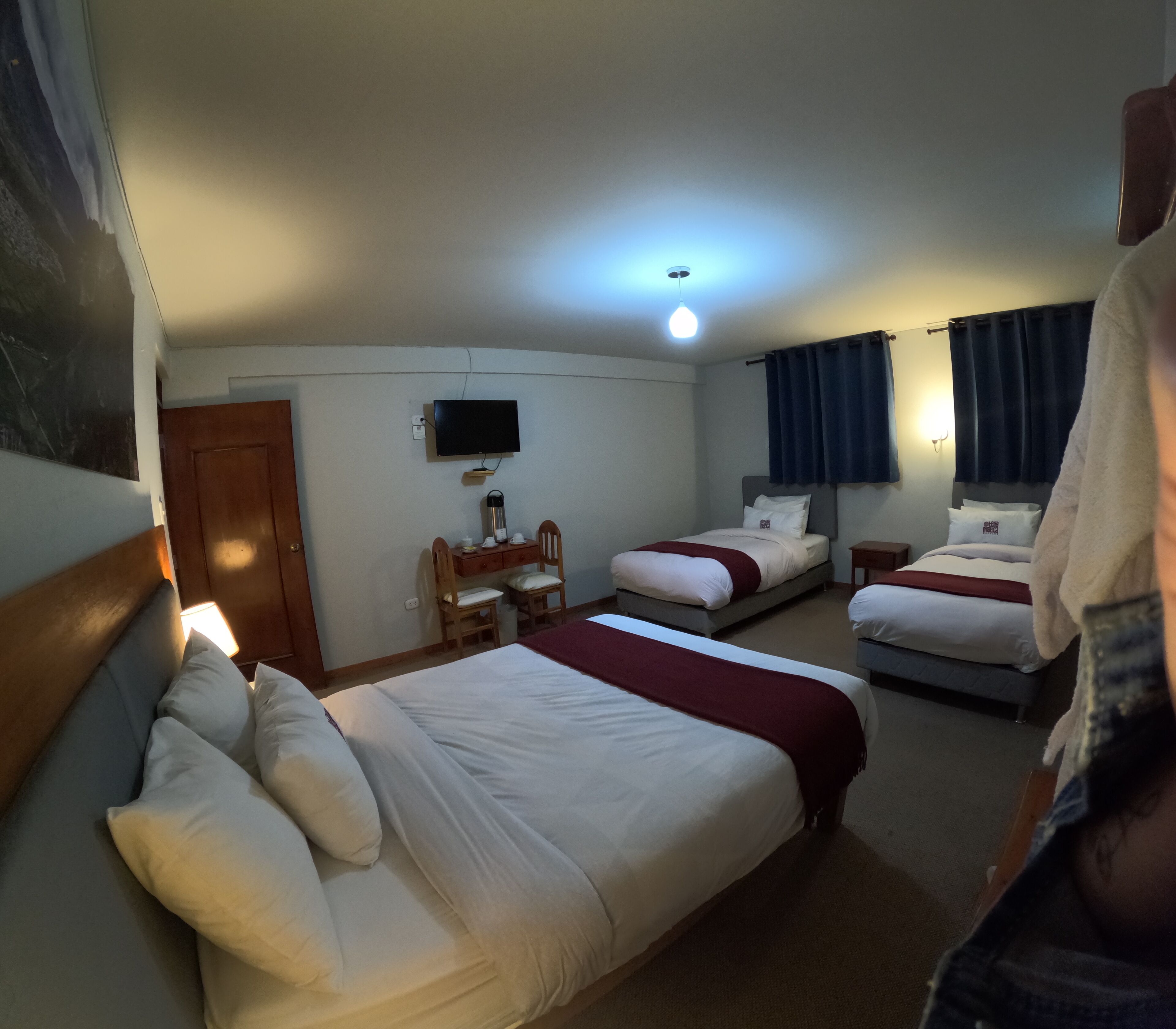 A hotel room with two beds, a desk, a chair, a TV, and a ceiling light.