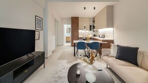 Una sala moderna con una televisión de pantalla plana, un sofá, una mesa de centro y un comedor con sillas, y una cocina al fondo.