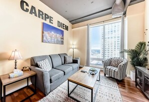 A living room with a grey sofa, a patterned armchair, a coffee table, and a large window with a city view. - 2 Full Beds, Memorable 1 Bedroom Condo, Downtown Central, Near Rogers Place (Edmonton)
