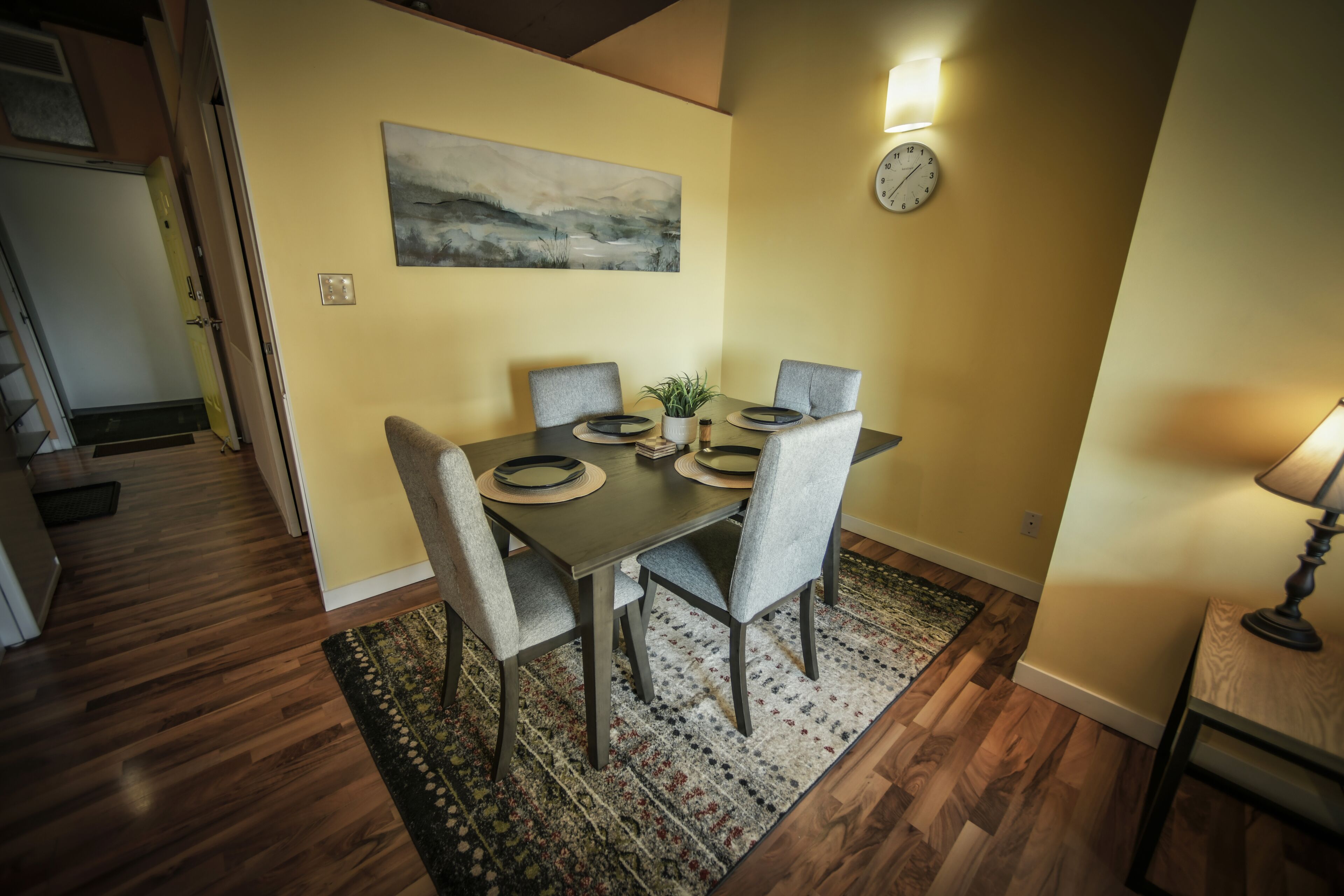A dining area with a table set for four, a painting on the wall, and a clock.