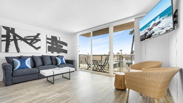 Una sala de estar moderna con un sofá gris, una mesa de centro y un sillón de mimbre. Se ve un ventanal con vistas al exterior.