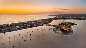 Vista aérea de un pueblo costero con un puerto deportivo lleno de barcos, una isla central con edificios y un atardecer sobre el agua.