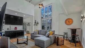Uma sala de estar moderna com um sofá cinzento, uma mesa de centro de vidro, uma televisão e uma grande janela com vista para edifícios.