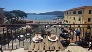 Een balkon met houten tafels, twee borden eten en uitzicht op een jachthaven met boten en een helderblauwe lucht.